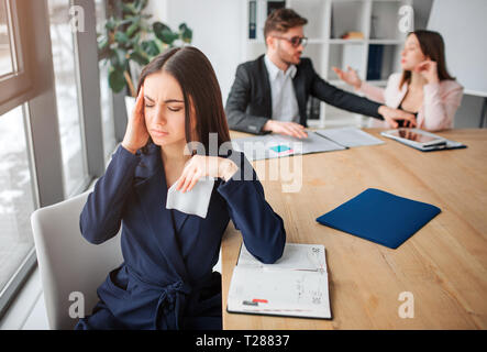 Kranke junge Frau am Tisch am Fenster sitzen. Sie halten Sie die Hand auf den Kopf und Leiden. Frau halten die Augen geschlossen. Ihr Partner arbeiten hinter Stockfoto