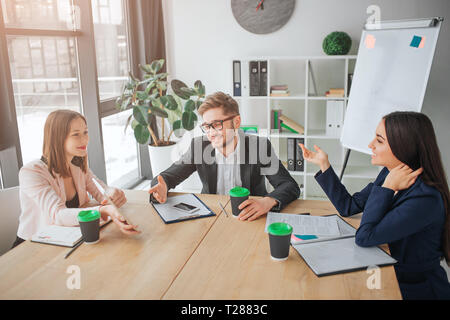Netter junger Mann und Frauen sitzen zusammen am Tisch im Konferenzraum. Sie haben Konversation und Lachen. Die Menschen arbeiten und sprechen Stockfoto