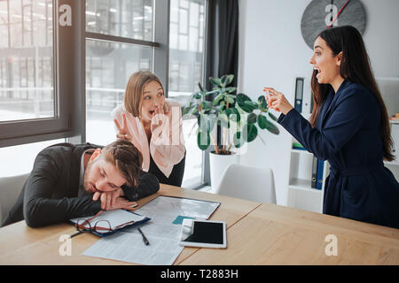 Müde junge Mann schlafen auf Tisch. Er halten Sie den Kopf auf die Hände. Lustig junge Frauen posieren und die Bilder mit ihm am Telefon Kamera. Sie lachen und Hav Stockfoto