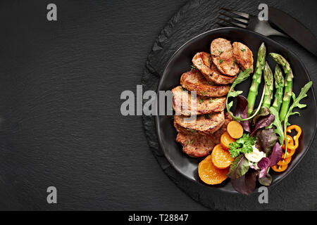 Blick von oben auf eine schwarze Platte mit gebratenen Scheiben Rindfleisch, Süßkartoffel und gemischter Salat auf schwarzem Schiefer Stockfoto