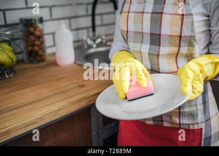 In der Nähe von Frau Hände in gelb Handschuhe Geschirr mit Schwamm. Sie stand in der Küche an der Spüle. Frau, die weiße Platte Stockfoto