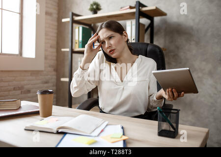 Junge busineswoman ist schwierig sprechen am Telefon. Sie sitzen am Tisch im Zimmer. Modell Tablet. Stockfoto