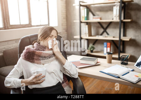 Kranke junge geschäftsfrau am Tisch im Zimmer sitzen und Niesen. Sie leiden an der Krankheit. Frau kann nicht arbeiten. Stockfoto