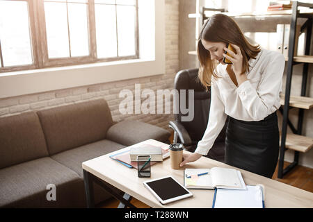Beschäftigte junge Frau stantd am Tisch im Zimmer. Sie reden über das Telefon und im Notizbuch schreiben. Stockfoto