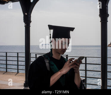 Männliche Kursteilnehmer, mit Graduierung Kleid, mit Smartphone, im Küstenort Stanley, Hong Kong Stockfoto