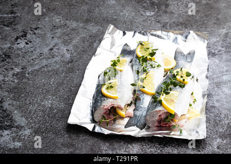Frische ganze Wolfsbarsch Fisch mit Zitrone und Kräutern auf Folie auf grauem Stein Tabelle Stockfoto