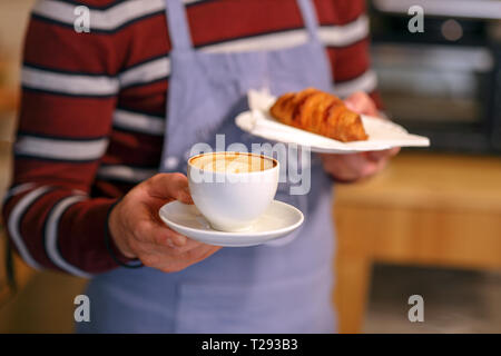 Barista mit Cappuccino und Croissant. Nahaufnahme auf der Hand des Mannes. Selektiver Fokus auf Cappuccino Stockfoto