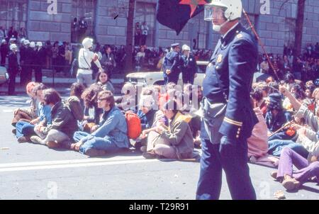 Während der 1971 Tag der Proteste gegen den Vietnamkrieg, einer Gruppe von Warm gekleidete männliche und weibliche Demonstranten sitzen zusammen in der Mitte des 10. ST NW, an einem sonnigen Tag, eine off-kamera Abbildung Wellen eine schwarze, rote und grüne Jugend internationale Partei' oder 'Yippie' Flag, in Washington, DC, Mai 1971. () Stockfoto