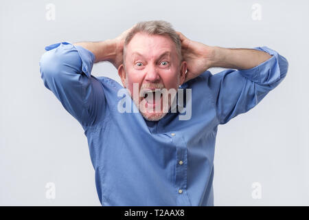 Älterer Mann im blauen Hemd mit wütenden Grimasse auf seinem Gesicht, mit dem Mund in Schreien geöffnet, bereit, zu argumentieren. Studio schießen. Negative menschliches Gefühl. Stockfoto