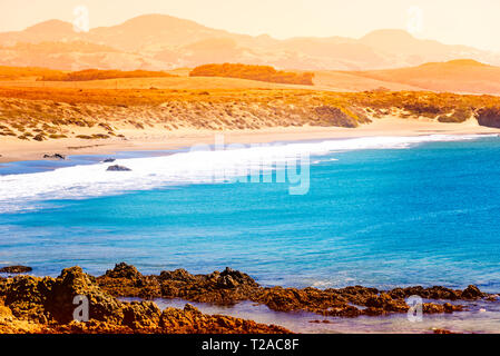 Mit Blick auf den felsigen Küstenlinie mit goldenen Sandstränden und dunstige Sommer Berge, helle blaue Meer mit weißen Wellen an den Stränden. Stockfoto