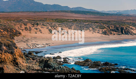 Mit Blick auf das blaue Meer mit Wellen auf die Felsen brechen und Strand unterhalb, Felder mit Dunstige Berge unter hellen Himmel. Stockfoto