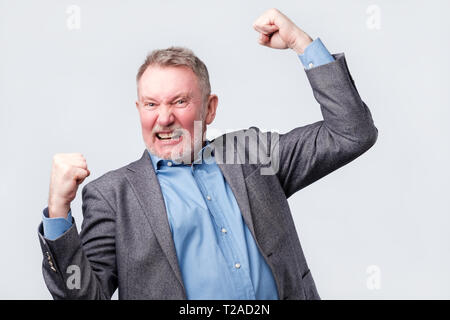 Gerne älterer stattlicher Mann in Anzug und blaues Hemd und Brille Gestik und halten den Mund. Er ist glücklich und feiert seinen Erfolg. Stockfoto