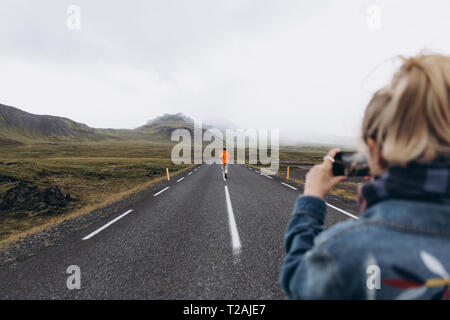 Junge Frau fotografieren Freund zu Fuß auf der Autobahn in Island Stockfoto