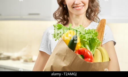 Vorderansicht des lächelnden jungen Frau mit weißem T-Shirt und die Papiertüte mit Essen an Küche. Froehlich holding Gemüse, Grün, Brot für c Stockfoto