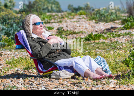 Ältere Frau im Mantel Sitzen auf einem Liegestuhl an einem kalten Tag im Frühjahr in Großbritannien. Stockfoto