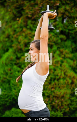 Lächelnd schwangere Frau tun Trizeps Training mit Hantel in Park Stockfoto