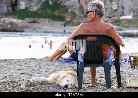 Toskana, Italien, 17. JULI 2018: reifer Mann ruht auf einem Liegestuhl Musik petting seinen Hund am Strand. Stockfoto