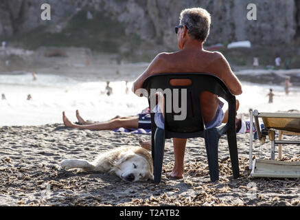 Toskana, Italien, 17. JULI 2018: reifer Mann ruht auf einem Liegestuhl Musik petting seinen Hund am Strand. Stockfoto