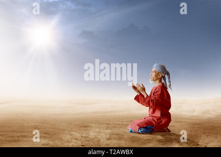 Asiatische muslimischen Mann mit Turban in seinem Kopf knien und beten mit erhobenen Händen auf Wüste mit Sonnenstrahlen und dunklen Himmel Hintergrund Stockfoto