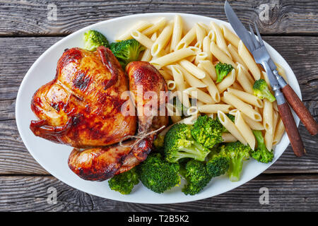 Ganzes gebackenes Huhn mit Brokkoli und pasta Penne Rigate auf einem weißen Teller mit Messer und Gabel, auf einem grauen rustikalen Holztisch, Ansicht von oben Stockfoto