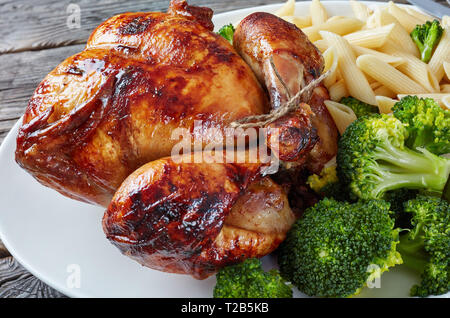 Ein ganzes gebratenes Huhn mit Brokkoli und Penne Rigate auf einem weißen Teller mit Messer und Gabel, auf einem grauen rustikalen Holztisch, Ansicht von oben, in der Nähe - u Stockfoto