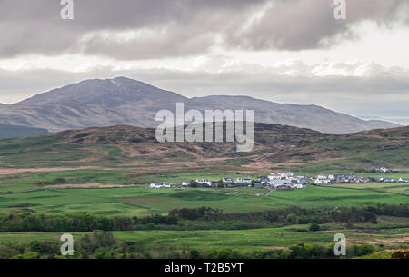 Ein kleines Dorf auf der Insel Jura sitzt unterhalb großer Hügel und ist von landwirtschaftlichen Flächen umgeben. Von der Insel Islay, Schottland gesehen. Stockfoto