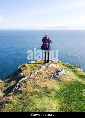 Ein Wanderer mit einem kleinen roten Rucksack steht auf einer Klippe über dem Meer an einem sonnigen Tag. An der Spitze der Halbinsel, auf Oa t fotografiert. Stockfoto