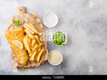 Traditionelle britische Fisch und Chips mit Sauce Tartar auf Schneidebrett und grünen Erbsen auf weißen Stein Hintergrund. Zitronenscheibe und Salz Stockfoto