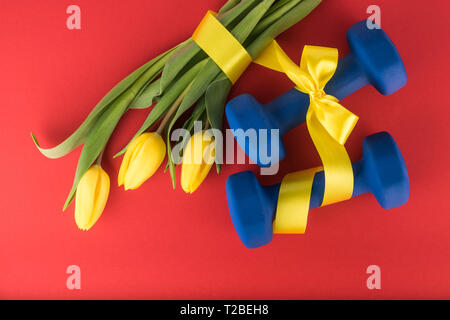 Drei gelbe Tulpen und Hanteln mit Bogen auf rotem Hintergrund Stockfoto