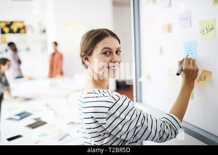 Taille bis Porträt des zeitgenössischen jungen Frau Zeichnung auf dem Whiteboard in Büro- und lächelnd an Kamera, kopieren Raum Stockfoto