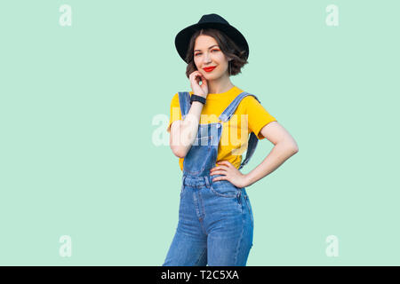 Beauty Portrait von hübschen jungen Hipster girl in Blue Jeans Overalls, gelbes Hemd, schwarzer Hut stehend mit der Hand an der Hüfte und in die Kamera lächeln Stockfoto