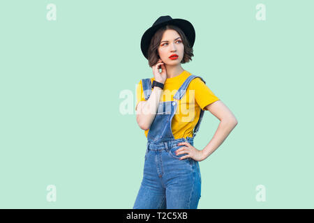 Portrait von nachdenklich junge Hipster girl in Blue Jeans Overalls, gelbes Hemd, schwarzer Hut stehend mit der Hand an der Hüfte, auf der Suche nach Pflege und Denken. Stockfoto