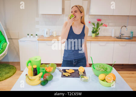 Junge lächelnde blonde Frau beißt Bio Apfel beim Kochen frische Früchte in der Küche. Das gesunde Essen. Vegetarische Mahlzeit. Diät Entgiftung Stockfoto