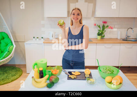 Junge lächelnde blonde Frau hält Gebissen grüner Apfel beim Kochen frische Früchte in der Küche. Das gesunde Essen. Vegetarische Mahlzeit. Diät Entgiftung Stockfoto