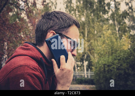 Schöner modischer Hipster in Sonnenbrille im Gespräch per Telefon auf dem Hintergrund der Herbst Park. junger Mann macht einen Anruf auf einem Mobiltelefon. Stockfoto