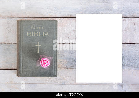 Heilige Bibel mit dem Wort biblia in Spanisch und ein Kreuz und eine Rose Blume Dekoration neben einem leeren weißen Papier auf einem Holztisch. Ansicht von oben und Fram Stockfoto