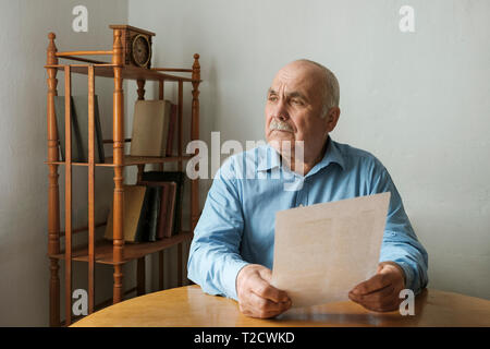 Mann sitzt der Betrachtung ein schriftliches Dokument, das er in seinen Händen hält mit Blick auf die Seite mit einem nachdenklichen Ausdruck, während an einem Tisch drinnen sitzt Stockfoto