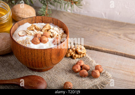 Hölzerne Schüssel mit Joghurt mit Haferflocken und Nüsse, Löffel, Glas mit Honig und Fir-Zweig auf sackleinen Serviette auf Holzplanken in der Nähe von weißen Wand. Selektive foc Stockfoto