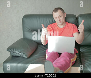 Lächelnder Mann mit Laptop sitzt auf einem Sofa zu Hause und hält Daumen hoch, blickt in die Kamera Stockfoto