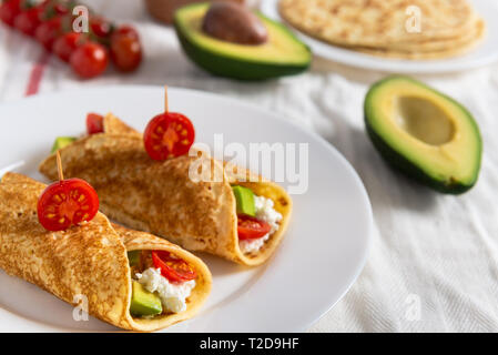 Crepe Pfannkuchen mit Avocado, weicher, weißer Käse und Kirschtomaten auf weiße Platte. Cherry Tomaten, Avocado und Stapel von Crepes auf der Seite. Stockfoto