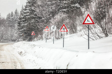 Zeile der Warnung Beschilderung auf gefährlichen Teil der Forststraße, im Winter Blizzard. Achtung Rehe, Schnee, Kufen und Steinschlag. Stockfoto