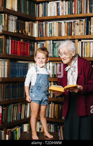 Glückliche Momente mit ihrer Urgroßmutter, ältere Dame viel Zeit mit ihrem großen Enkelin. Portrait vor einem Bücherregal, ein Buch, das ich Stockfoto