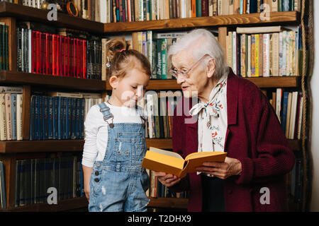 Glückliche Momente mit ihrer Urgroßmutter, ältere Dame viel Zeit mit ihrem großen Enkelin. Portrait vor einem Bücherregal, sprechen über Buch Stockfoto