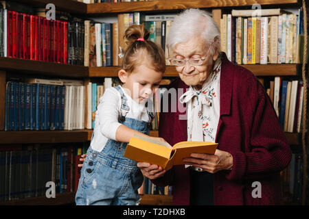 Glückliche Momente mit ihrer Urgroßmutter, ältere Dame viel Zeit mit ihrem großen Enkelin. Portrait vor einem Bücherregal, ein Buch, das ich Stockfoto