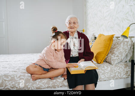 Glückliche Momente mit ihrer Urgroßmutter, ältere Dame viel Zeit mit ihrem großen Enkelin. Das Sitzen auf einem Bett, zusammen Buch zu lesen. Stockfoto