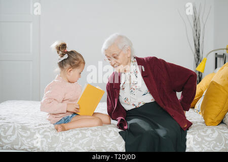 Glückliche Momente mit ihrer Urgroßmutter, ältere Dame viel Zeit mit ihrem großen Enkelin. Beide sitzen auf einem Bett, Mädchen lesen Buch und Gran Stockfoto