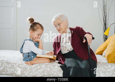 Glückliche Momente mit ihrer Urgroßmutter, ältere Dame viel Zeit mit ihrem großen Enkelin. Beide sitzen auf einem Bett, Mädchen lesen Buch und Gran Stockfoto