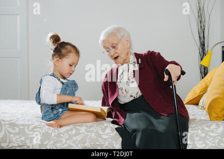 Glückliche Momente mit ihrer Urgroßmutter, ältere Dame viel Zeit mit ihrem großen Enkelin. Beide sitzen auf einem Bett, Mädchen lesen Buch und Gran Stockfoto