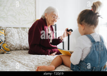 Glückliche Momente mit ihrer Urgroßmutter, ältere Dame viel Zeit mit ihrem großen Enkelin. Beide sitzen auf einem Bett, Mädchen lesen Buch und Gran Stockfoto