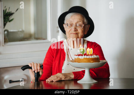 Respektable gut in Schwarz mini-hut grau behaarte ältere Frau mit Zuckerrohr ihren 90. Geburtstag bei Tisch zu Hause ist feiern gekleidet. Stockfoto